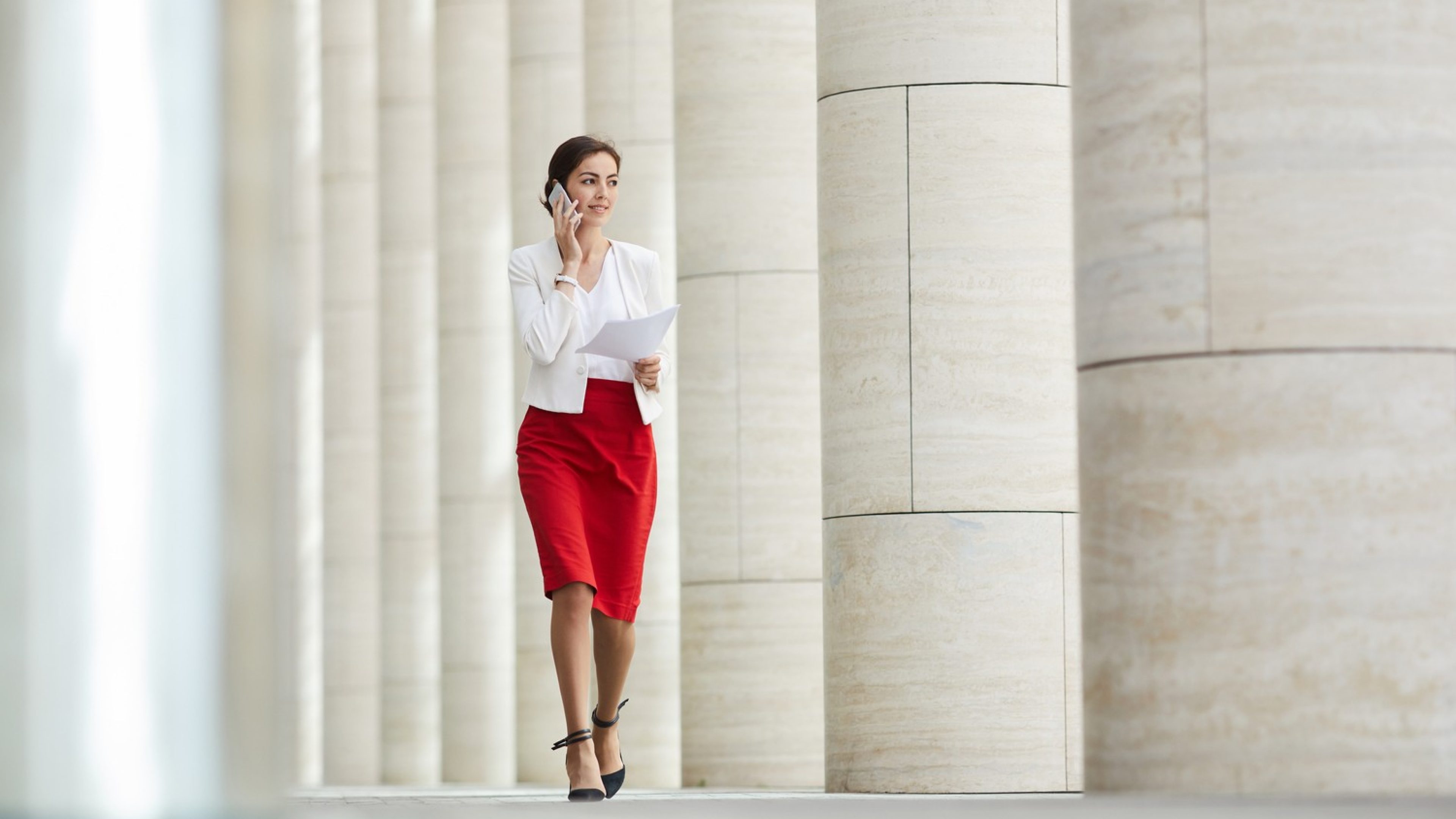 Full length portrait of beautiful woman speaking by phone while walking towards camera along row of pillars, copy space