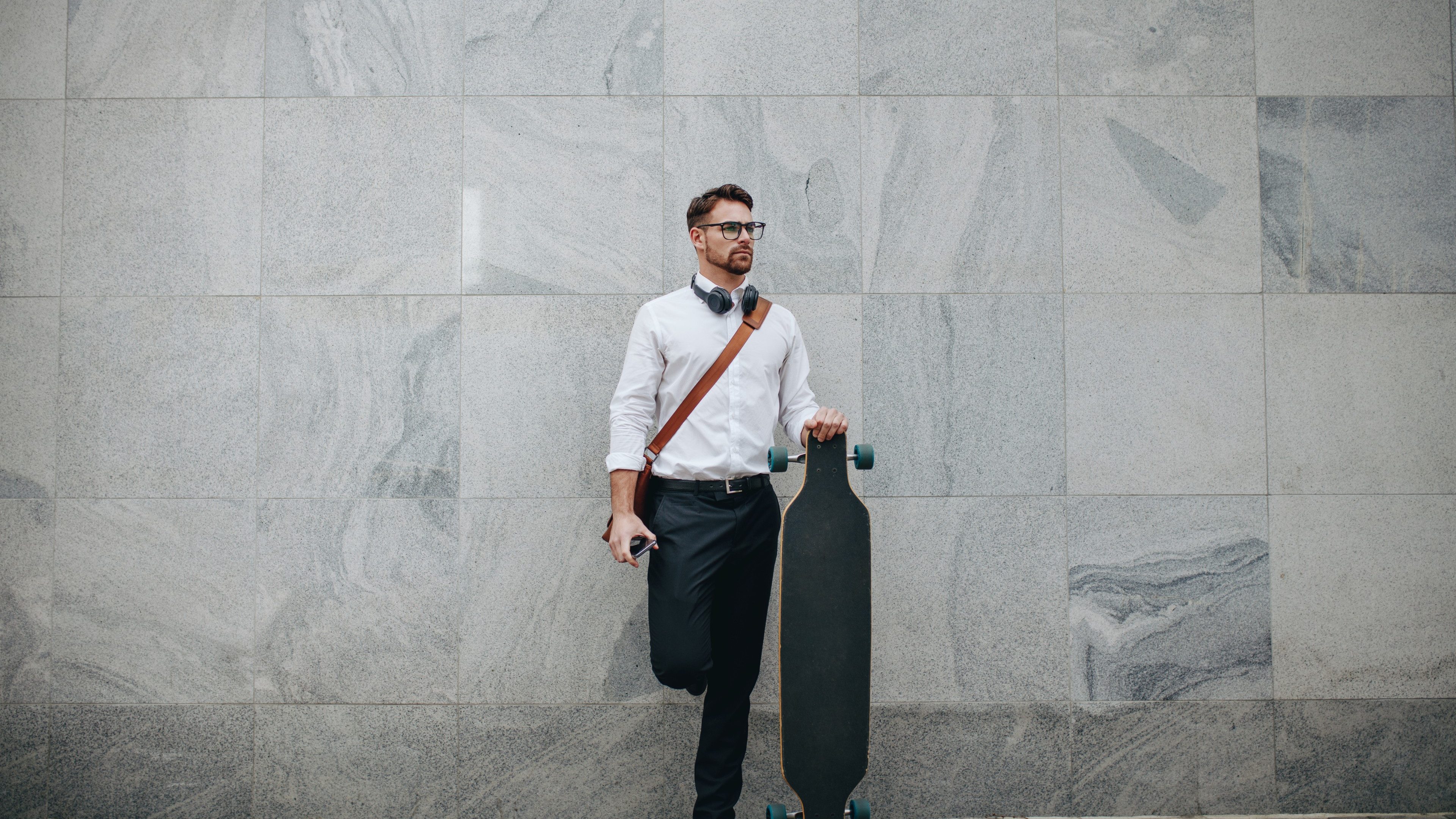 Formally dressed man standing in street with a longboard. Businessman standing against a wall outdoors holding a long skateboard.