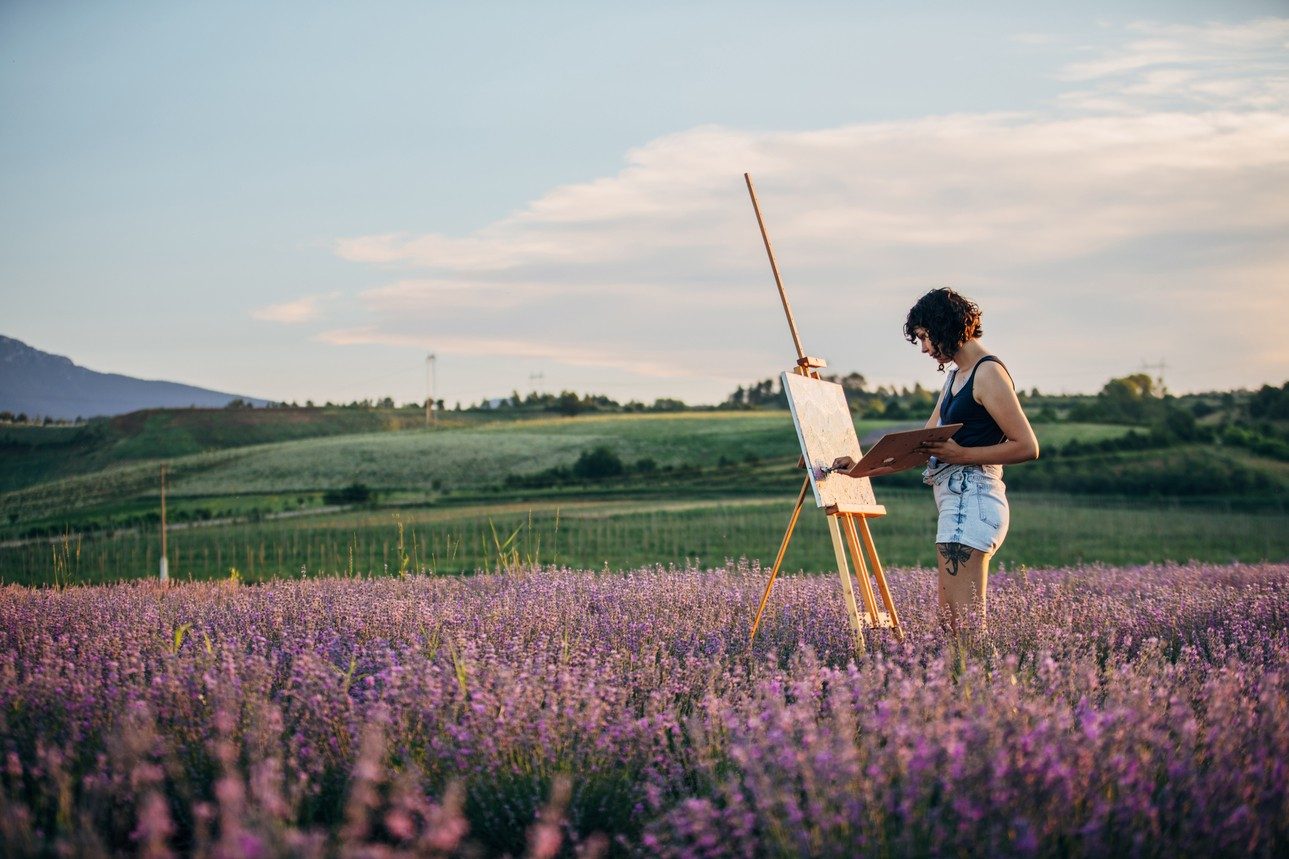 One woman, beautiful female painter artist painting on lavender field outdoors on a sunny day.