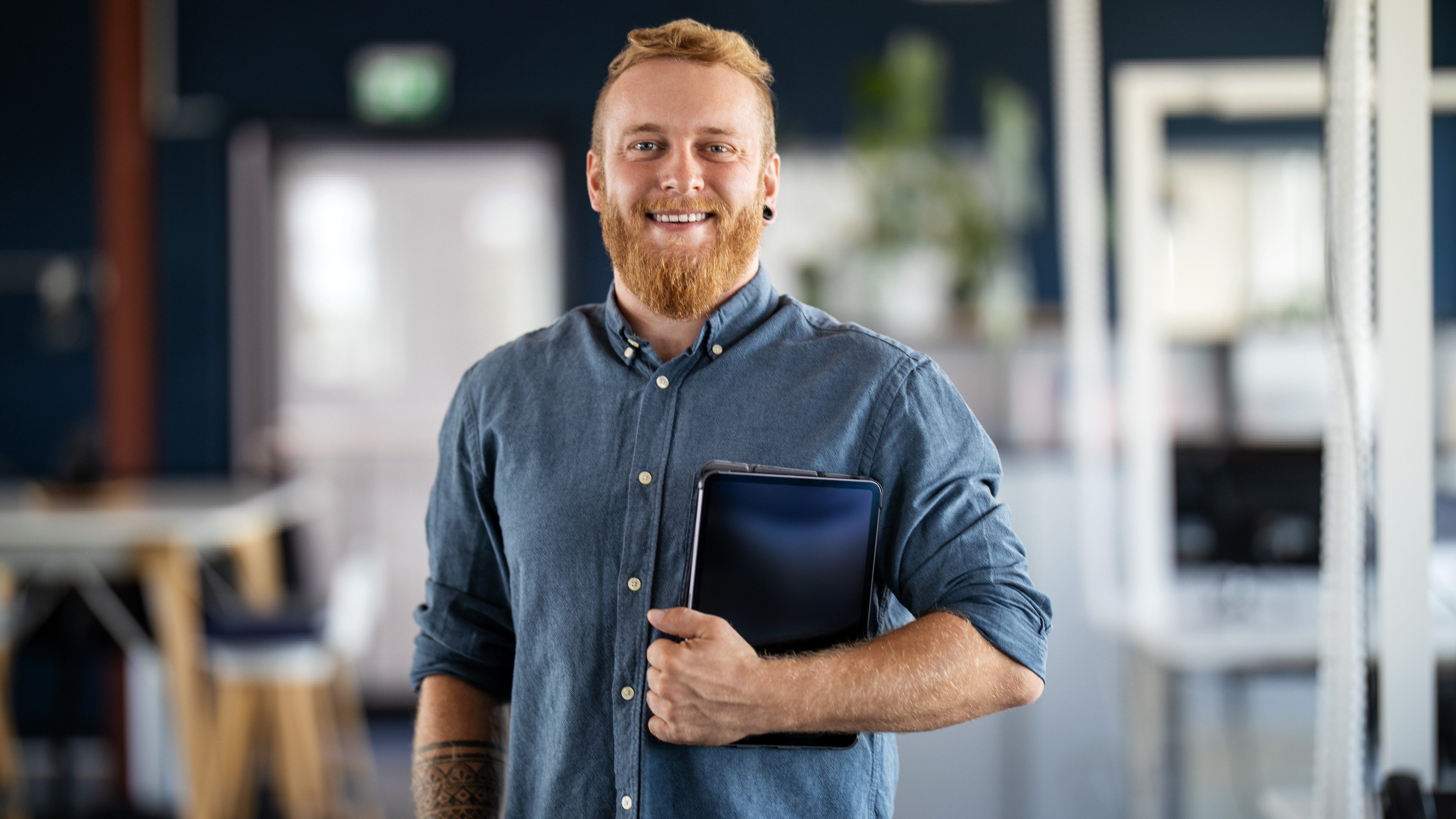 Portrait of a businessman with beard standing in office holding digital tablet. Confident male business executive in office looking at camera.
