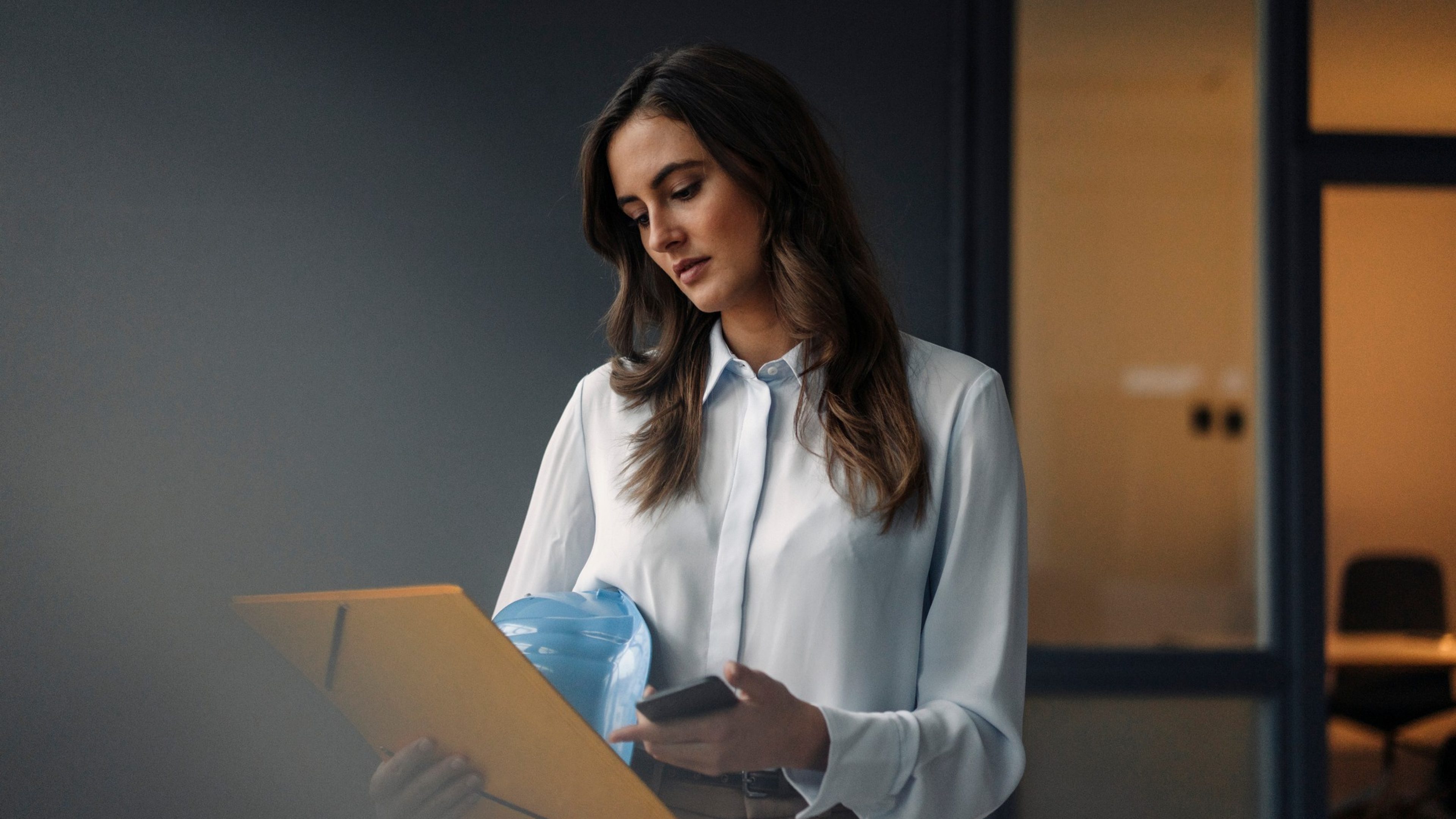 Serious young businesswoman holding folder, hard hat and cell phone